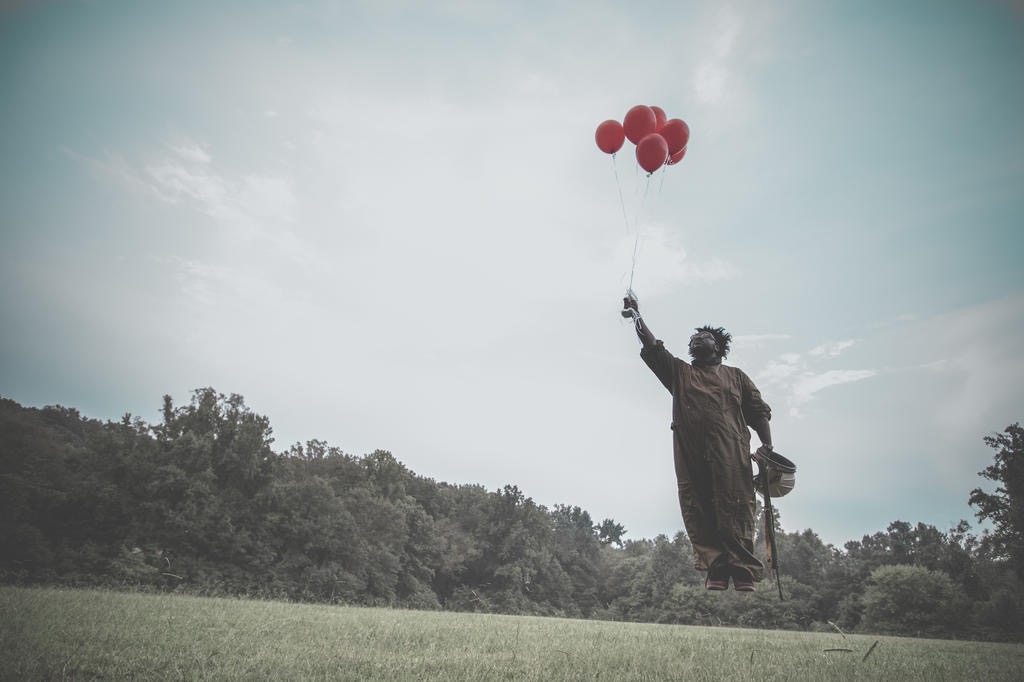 Figure carried aloft by red balloons above tall grass